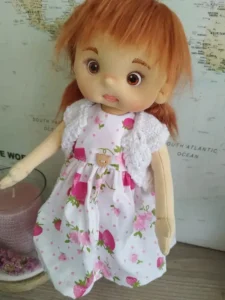 Mu&ntilde;eca de tela articulada soft de 37 cm con vestido de fresas, pelo rojizo y rebeca blanca.