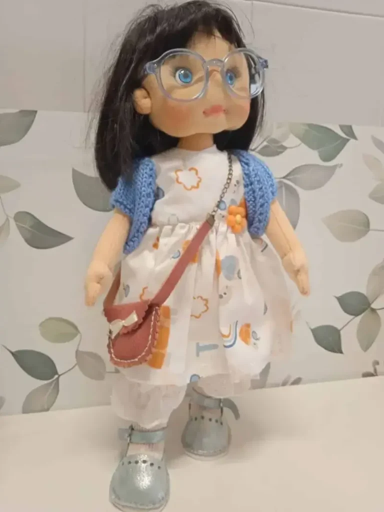 Mu&ntilde;eca de tela artesanal hecha a mano con pelo negro, gafas, rebeca de ganchillo azul, mini bandolera marr&oacute;n y zapatos plateados.