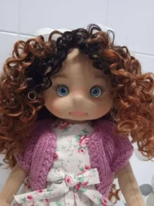 Plano medio de la mu&ntilde;eca de trapo artesanal destacando su melena de pelo rizado moreno y sus ojos azules.