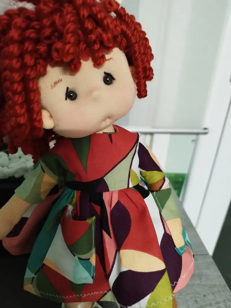 Muñeca de tela artesana Maryxeniss: muñeca con pelo de lana granate en tirabuzones, ojos negros y vestido geométrico de colores.