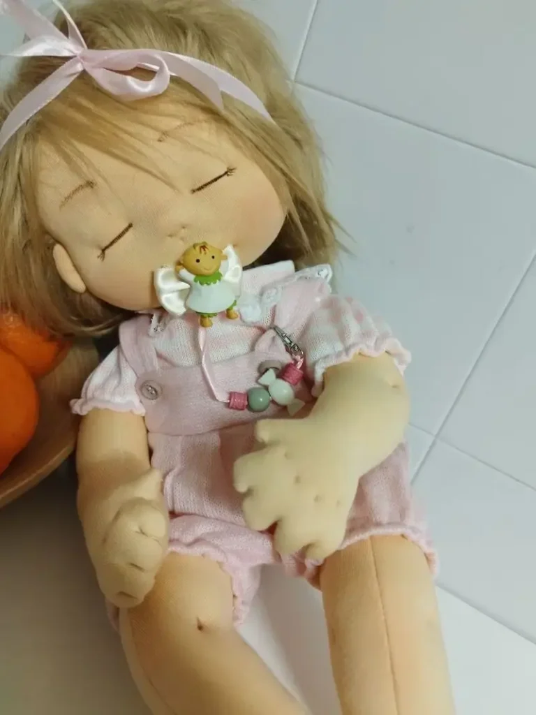 Mu&ntilde;eca de tela artesana Maryxeniss en versi&oacute;n beb&eacute;: mu&ntilde;eca de pelo rubio con ojos cerrados, lazo de sat&eacute;n y chupete.